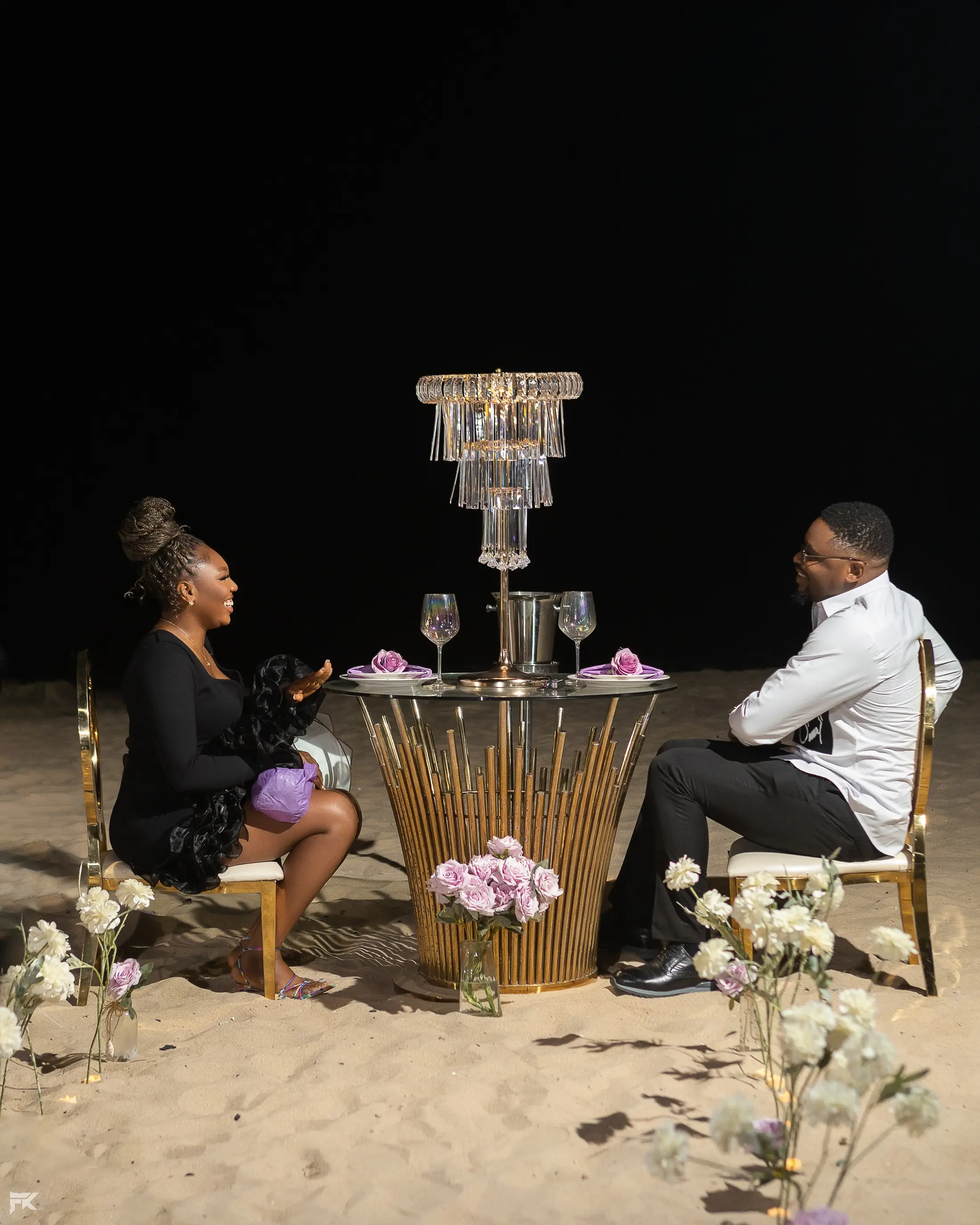 A couple dining at a candlelit table on the sand at night under a crystal chandelier