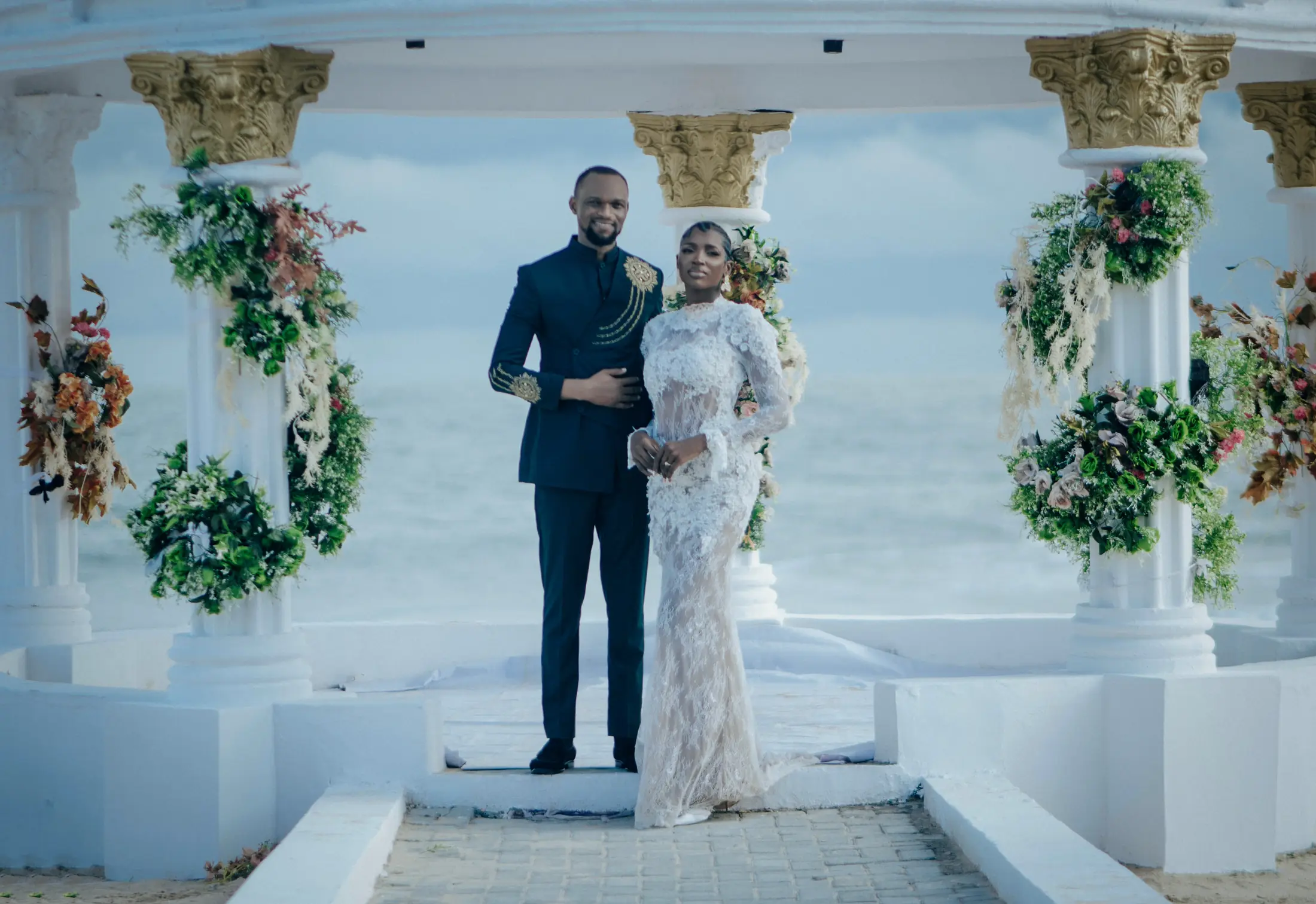 A couple standing together at the flower-dressed gazebo, the sea behind